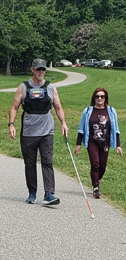 Front view image of blind male hiker wearing VibroGuide vest walking on a paved surface and also using white cane. Girl walking next to him is chatting.